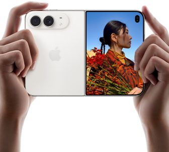 iPhone Duo, couleur blanc stellaire, deux mains le tiennent en orientation horizontale, déplié, le volet gauche montre les deux caméras arrière Fusion 48 mégapixels, le volet droit montre l’écran externe affichant une personne en train de se faire photographier, qui peut se voir pendant la prise grâce à Duo Preview